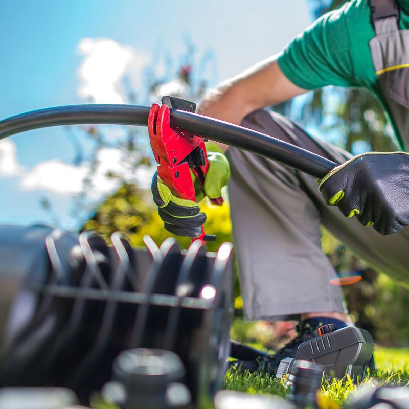 Person using a red pipe cutter on a black plastic pipe outdoors, wearing gloves and green shirt, with tools nearby.