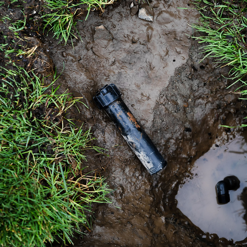 Exposed broken irrigation pipe in muddy ground next to a small puddle, surrounded by patches of grass.