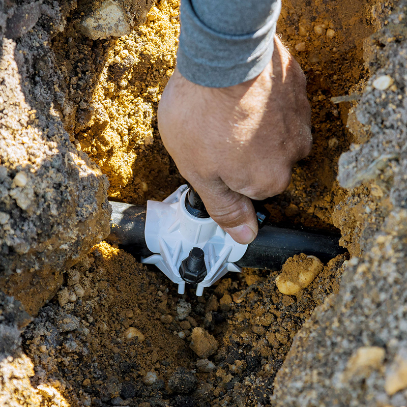 A person fixes a black plastic pipe using a white clamp in a trench surrounded by soil.