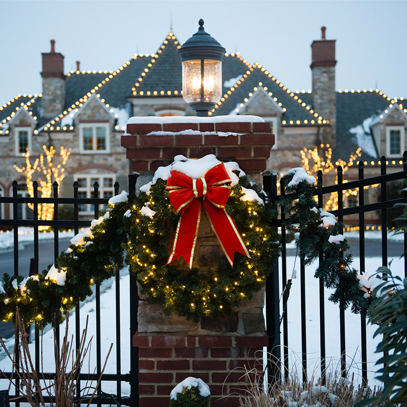 A snow-covered brick pillar with a festive wreath and red bow stands in front of a large house adorned with holiday lights.