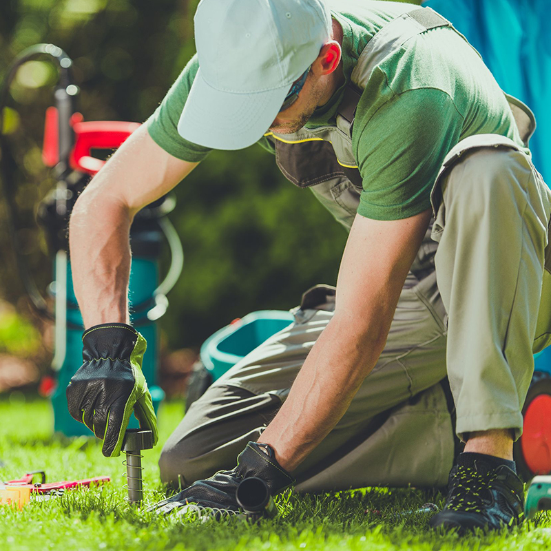 Person in gloves and cap kneeling on grass, adjusting a garden hose connector.