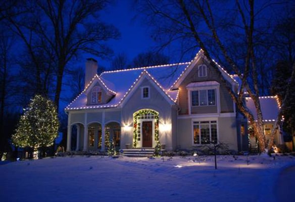 House at night with Christmas lights on roof and trees, surrounded by snow, under a clear dark blue sky.