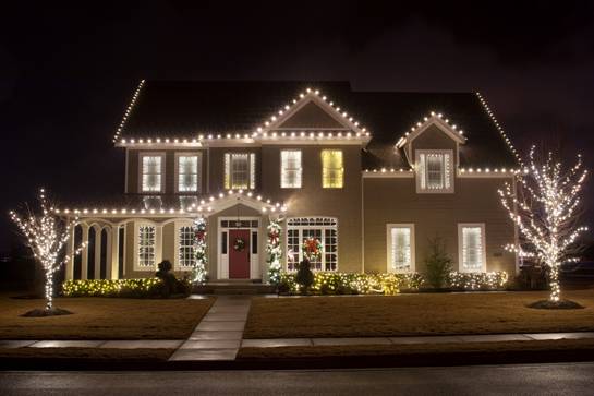 A two-story house is adorned with white Christmas lights, featuring illuminated trees and wreaths on the front door and windows.