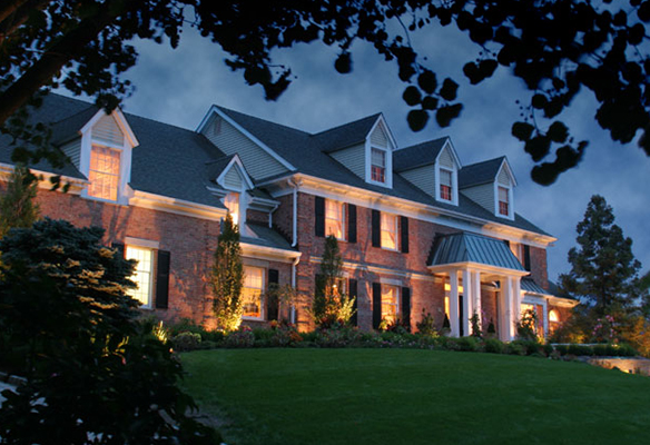 Large brick house at dusk with illuminated windows and well-kept lawn, framed by trees.