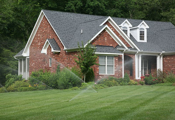 A red brick house with a manicured lawn and working sprinkler system, surrounded by trees.